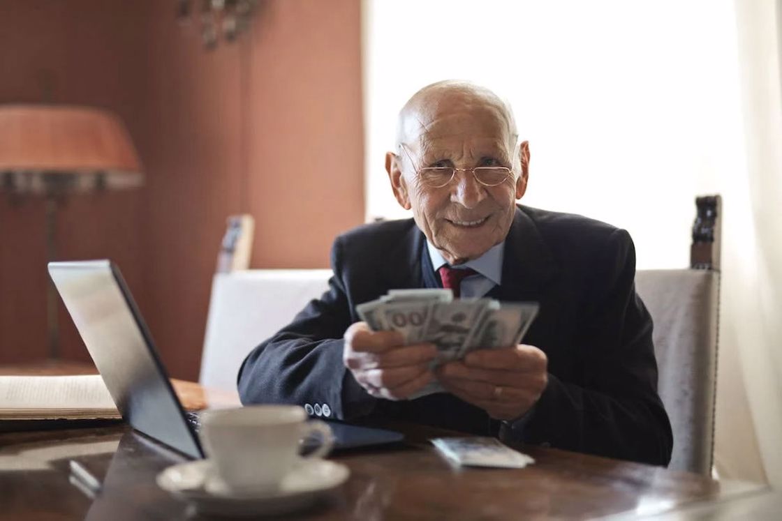 Senior businessman holding money in hands while sitting at table near laptop